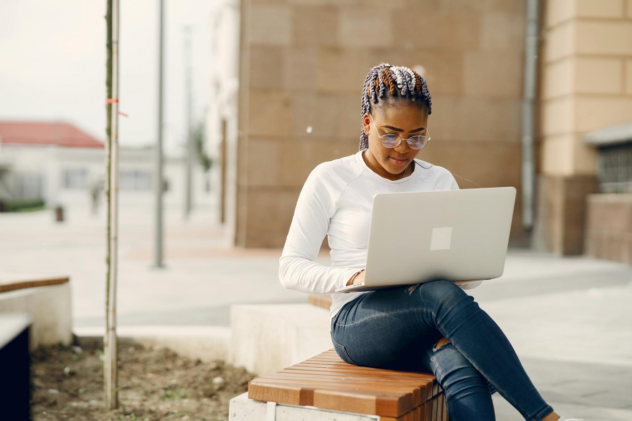 Young woman sitting on a bench, working on a laptop outdoors, enjoying a sunny day.